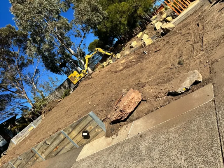 Excavator working beside sleeper stairs and a rock retaining wall on a suburban site